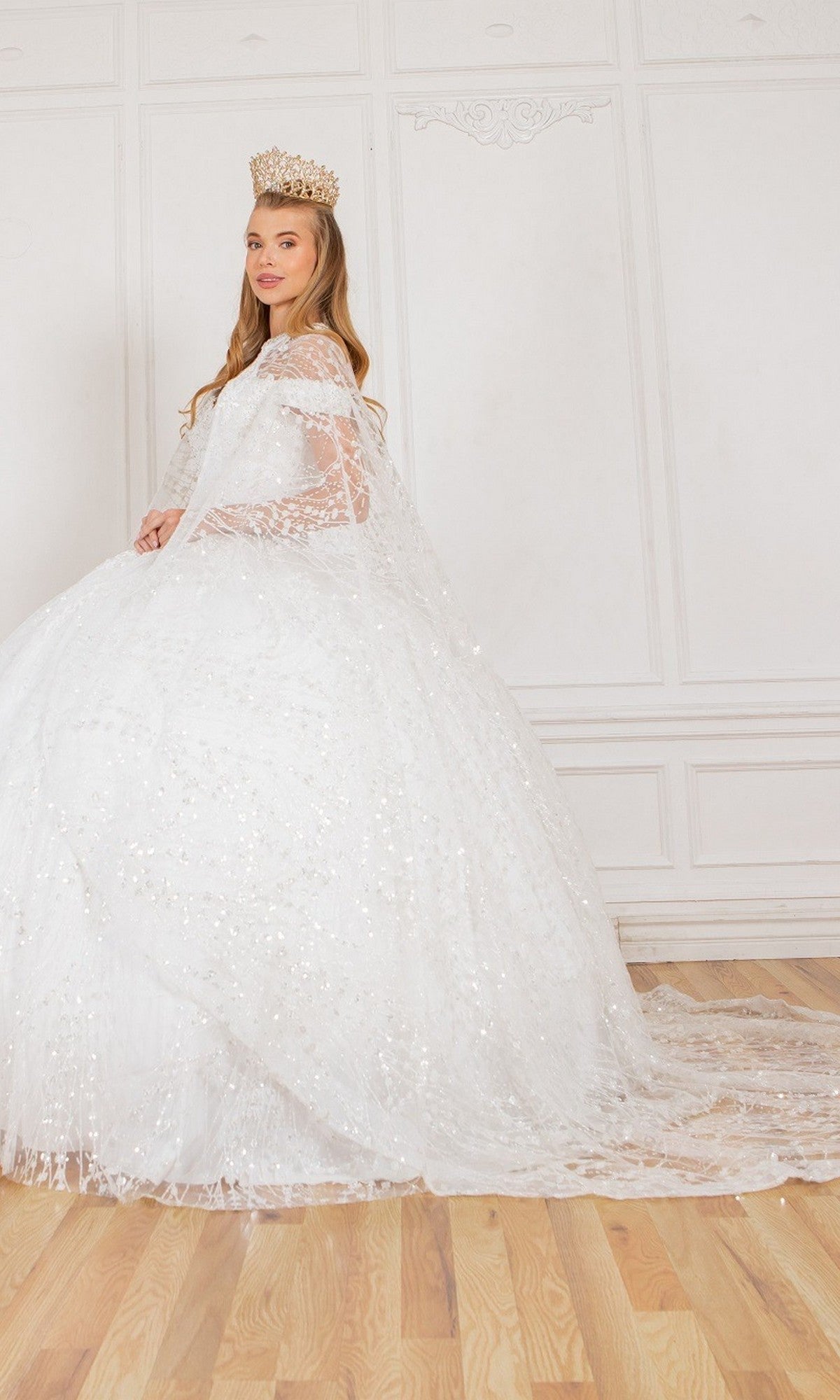 A woman stands indoors on a wooden floor, wearing the Quinceanera Dress 4201 by Cinderella Couture—a white, sparkling glitter ball gown with a matching cape—paired with a gold crown, set against a white paneled wall.